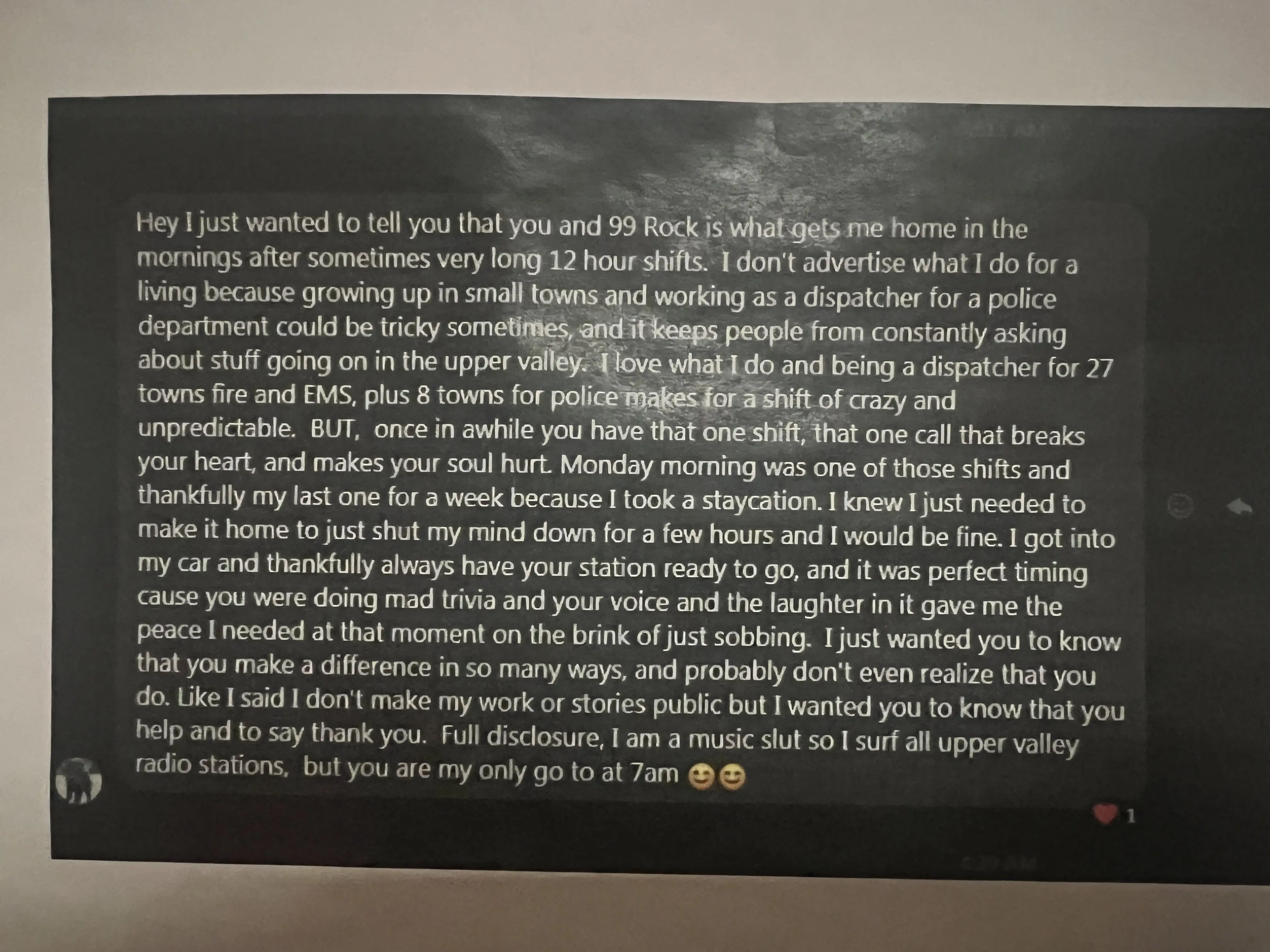 A printed out screenshot of a DM in darkmode against a white wall that reads, 
        &ldquo;Hey I just wanted to tell you that you and 99 Rock is what gets me home in the mornings after sometimes very long 12 hour shifts. 
        I don't advertise what I do for a living because growing up in small towns 
        and working as a dispatcher for a police department could be tricky sometimes, 
        and it keeps people from constantly asking about stuff going on in the upper valley. 
        I love what I do and being a dispatcher for 27 towns fire adn EMS, plus 8 towns for police makes for a shift of crazy and unexpected. 
        BUT, once in awhile you have that one shift, that one call that breaks your heart, and makes your soul hurt. 
        Monday morning was one of those shiftsadn thankfully my last one for a week because I took a staycation. 
        I knew I just needed to make it home to just shut my mind down for a few hours and I would be fine. 
        I got into my car and thankfully always have your station ready to go, 
        and it was perfect timing because you were doing mad trivia 
        and your voice and the laughter in it gave me the peace I needed in that moment on the brink of just sobbing. 
        I just want you to know that you make a difference in so many ways, and probably don't even realize that you do. 
        Like I said I don't make my work or stories public but I wanted you to know that you help and to say thank you. 
        Full disclosure, I am a music slut so I surf all upper valley radio stations, but you are my only go to at 7AM.&rdquo;