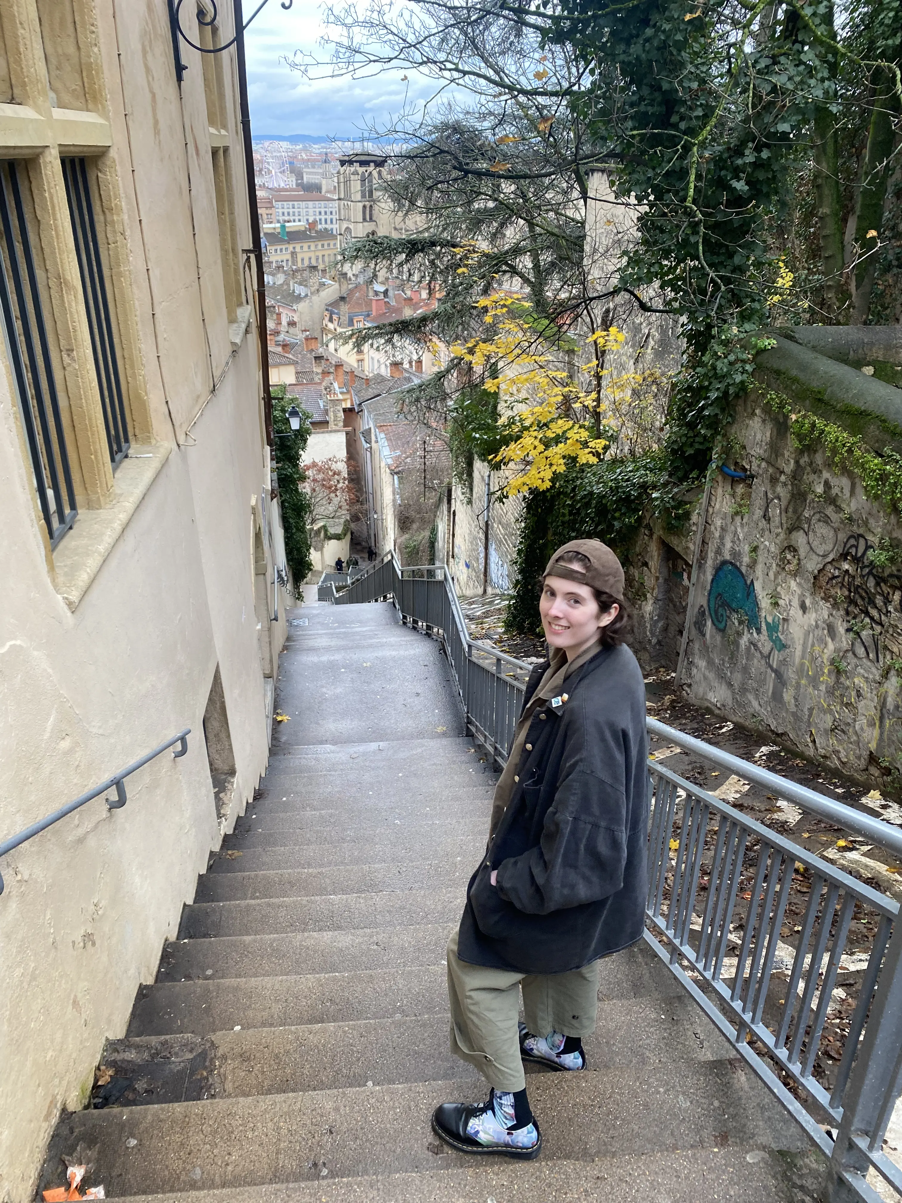 V smiles back at the camera warmly while standing on an outdoor staircase leading to an old city below. 
        You can see the beautiful, old city for miles from this high angle, 
        and yellow and green leaves, green ivy, and bare branches partially frame the background.
